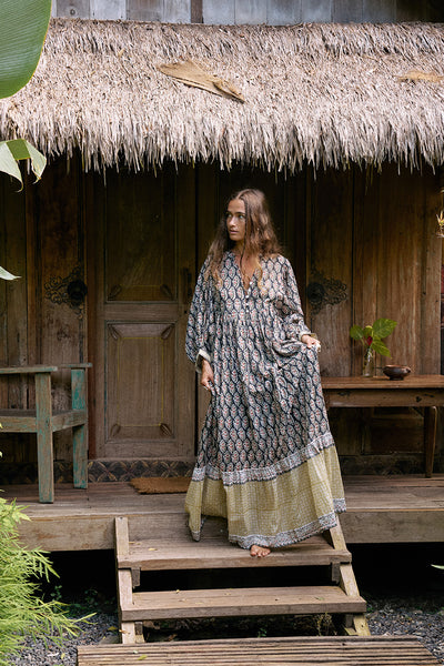 Bhoomi Maxi Dress Onyx — full length standing on wooden steps of thatched-roof hut, onyx hand block print maxi dress with contrast tiered hem, carved wooden door and tropical foliage | Daughters of India