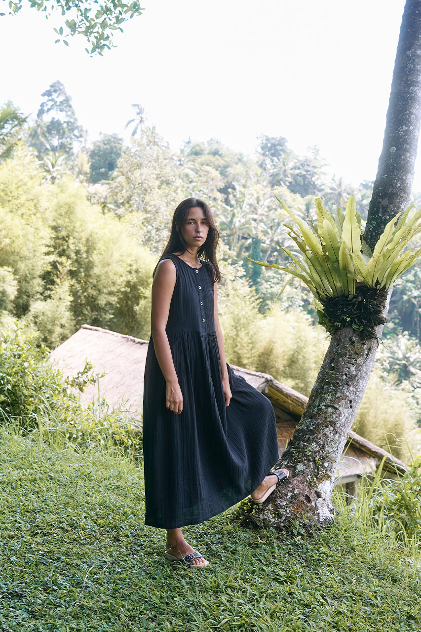 Nadi Dress Black — side view walking in tropical setting, black double gauze cotton midi dress showing flowing gathered skirt in motion | Daughters of India