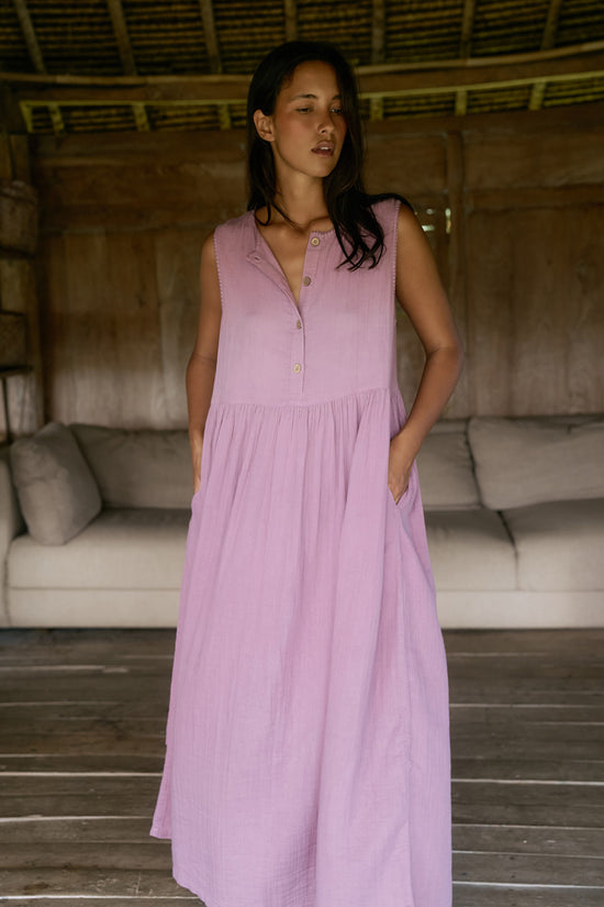 Nadi Dress Mauve — full length front view with hand in pocket, mauve double gauze cotton midi dress with wooden button front and gathered waist, rustic bamboo interior with sofa | Daughters of India