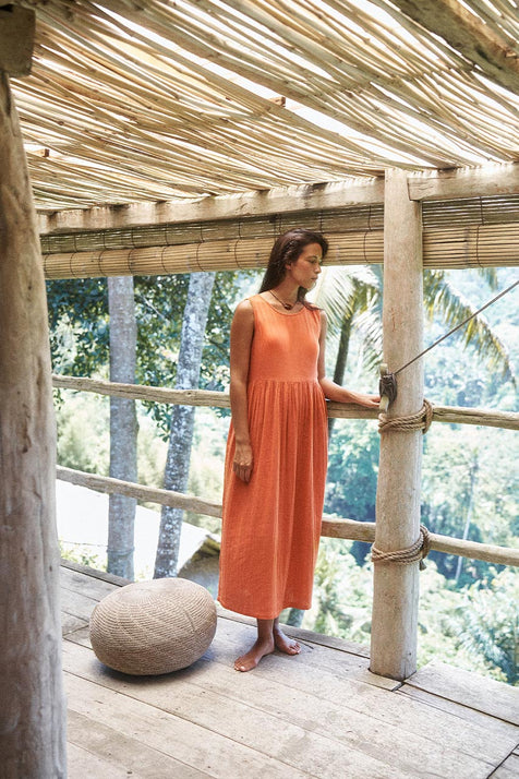 Nadi Dress Terracotta — full length side view on tropical bamboo veranda with palm trees, terracotta double gauze cotton midi dress with round neckline and gathered waist, rattan pouf | Daughters of India