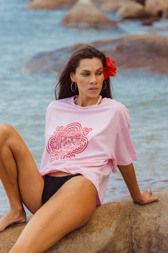 Shanti Tee Pink — seated on beach rocks, pink oversized cotton tee with red mandala logo, ocean and rocks backdrop, tropical coastal India | Daughters of India