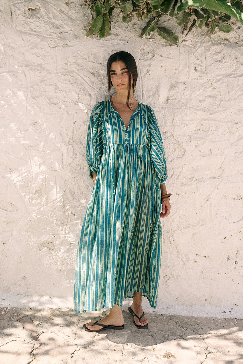 Full-length front view of woman wearing the Daughters of India Kyra Maxi Dress in Spearmint against a white wall in Greece, showing the flowing green striped silhouette